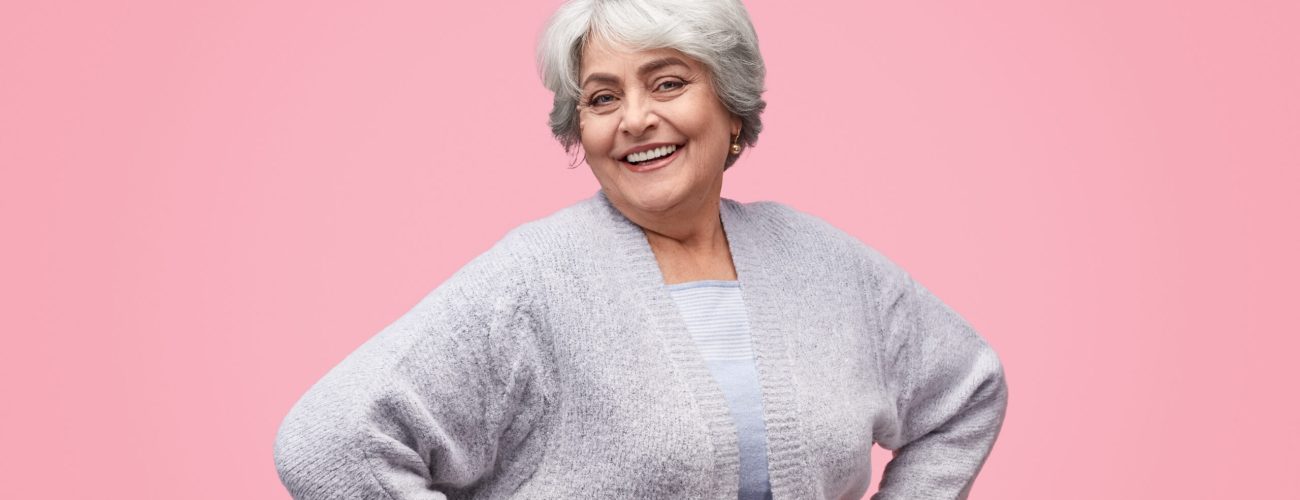 Young at heart elderly woman in elegant outfit keeping hands on waist and cheerfully smiling while standing on pink background and looking at camera